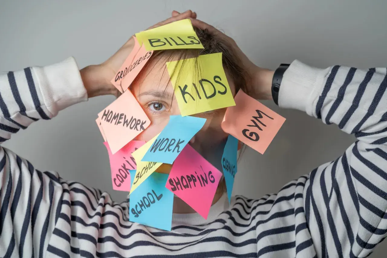 Una persona agobiada por notas adhesivas con tareas: "Kids", "Bills", "Homework", "Work", "Shopping", "Gym", "School", "Groceries", "Cleaning". Un recordatorio visual de cómo ser mejor persona es priorizar.