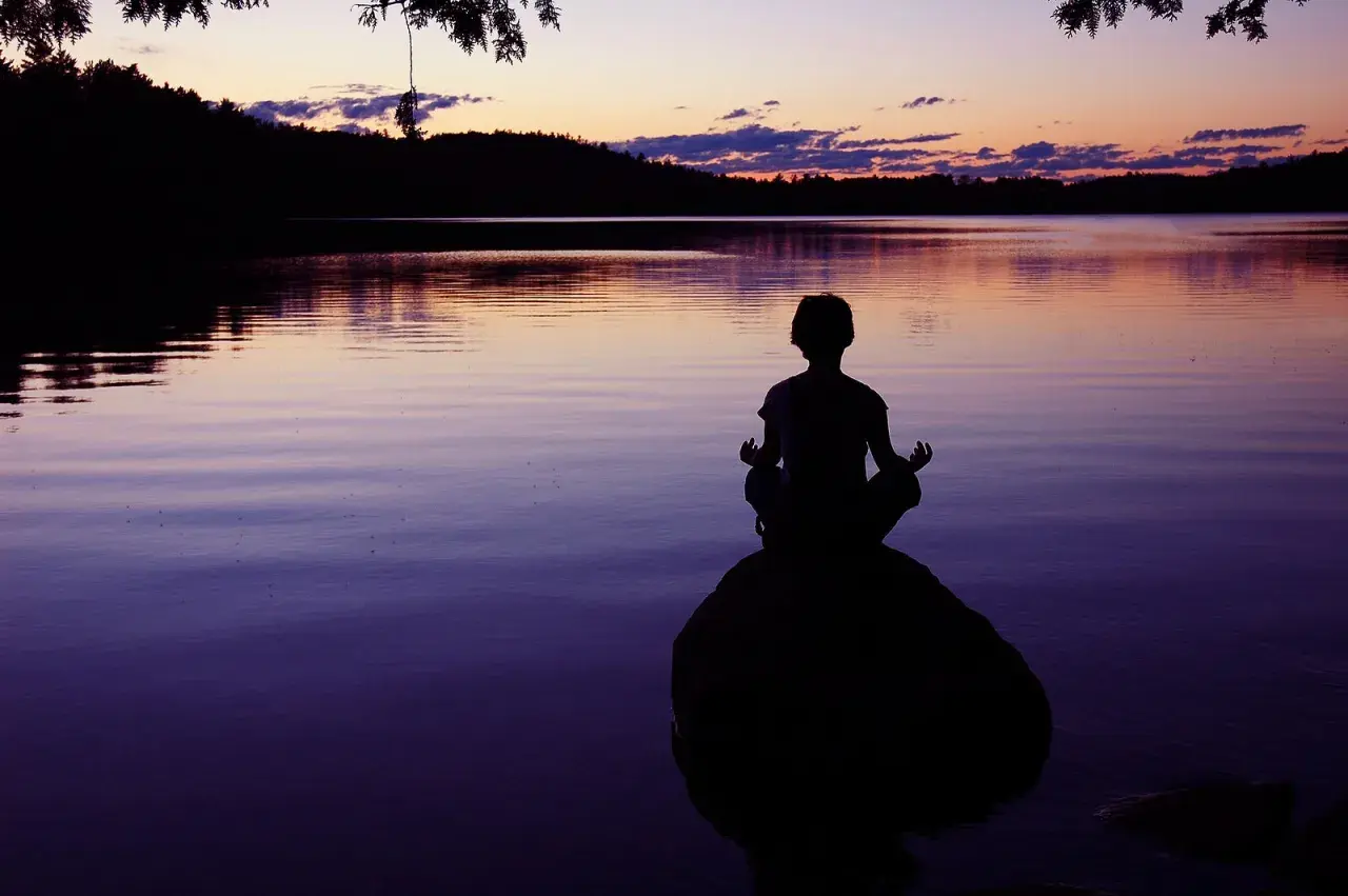 Silueta de persona meditando en una roca al atardecer, buscando la paz interior junto a un lago sereno.