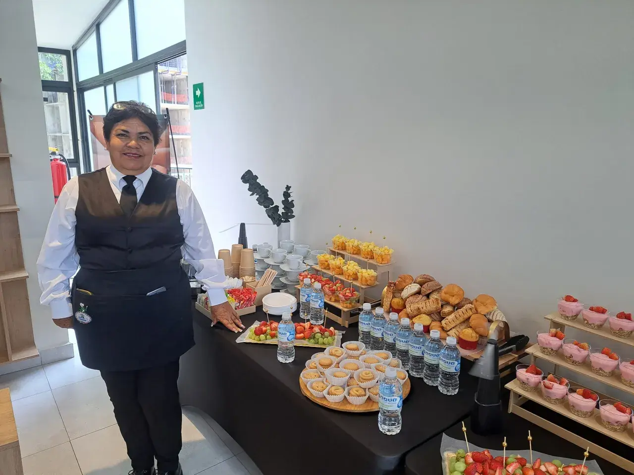 Una mujer sonriente supervisa un montaje de coffee break con frutas, postres, pan y agua.