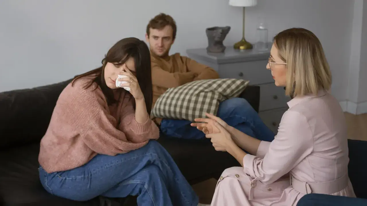 Una pareja discute temas de conversación difíciles con un terapeuta. La mujer llora mientras el hombre la mira.