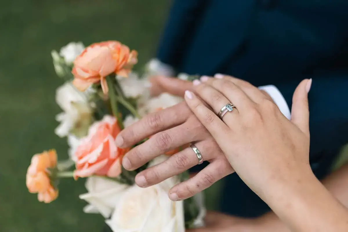 Manos entrelazadas con anillos, celebrando aniversarios de boda junto a un ramo de flores color melocotón y blanco.