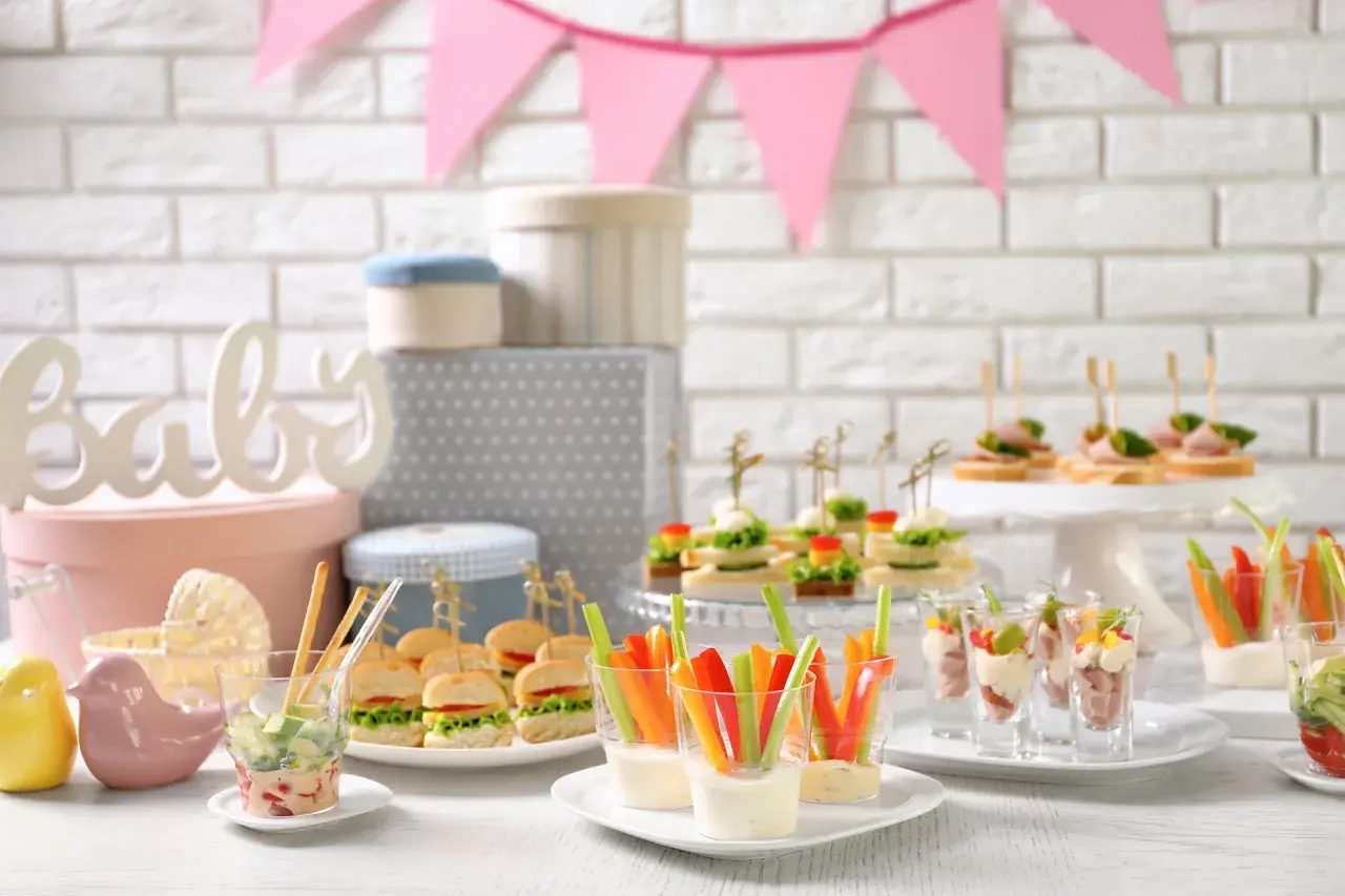Mesa de baby shower con deliciosa comida: mini hamburguesas, canapés, palitos de verduras con dip y vasitos de aperitivos.