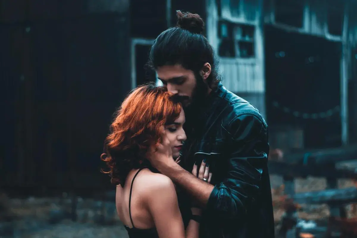 Un hombre besa la frente de una mujer con cabello rojizo. La escena evoca la complejidad de la infidelidad emocional, un vínculo secreto que florece en la sombra.