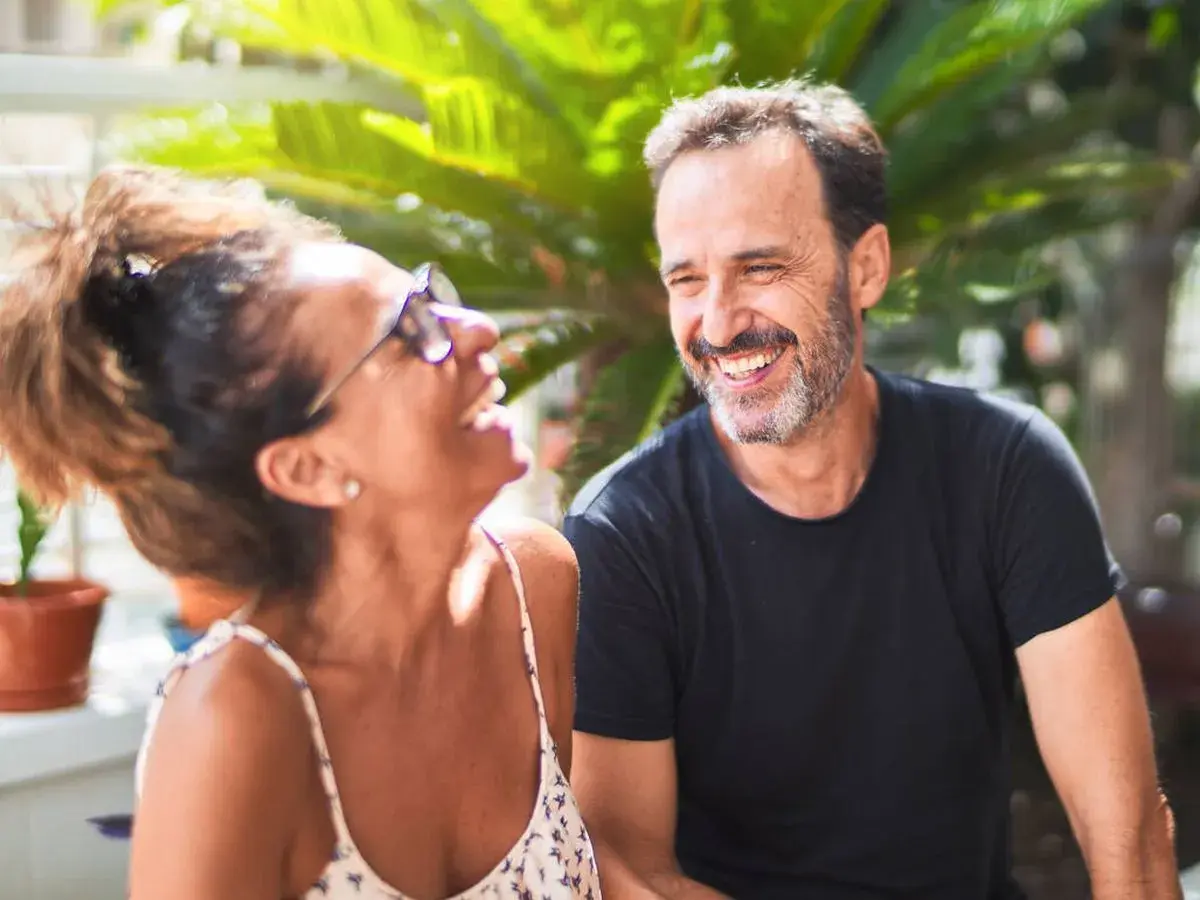 Pareja riendo al aire libre. Él, superando la crisis de los 50, sonríe con complicidad. Ella, con gafas de sol, se ríe a carcajadas.
