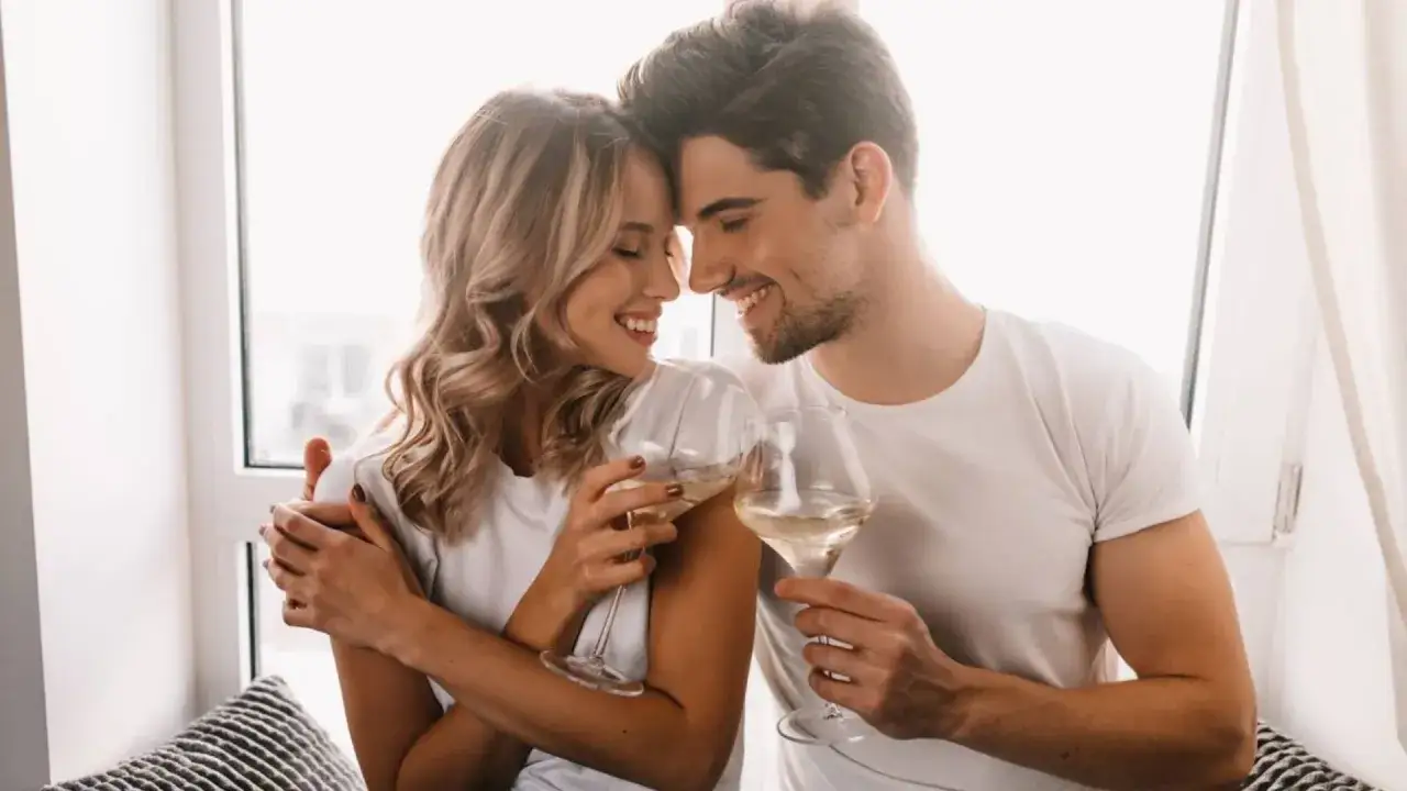 Pareja sonriente brindando con copas de vino blanco, celebrando sus aniversarios de boda junto a una ventana.