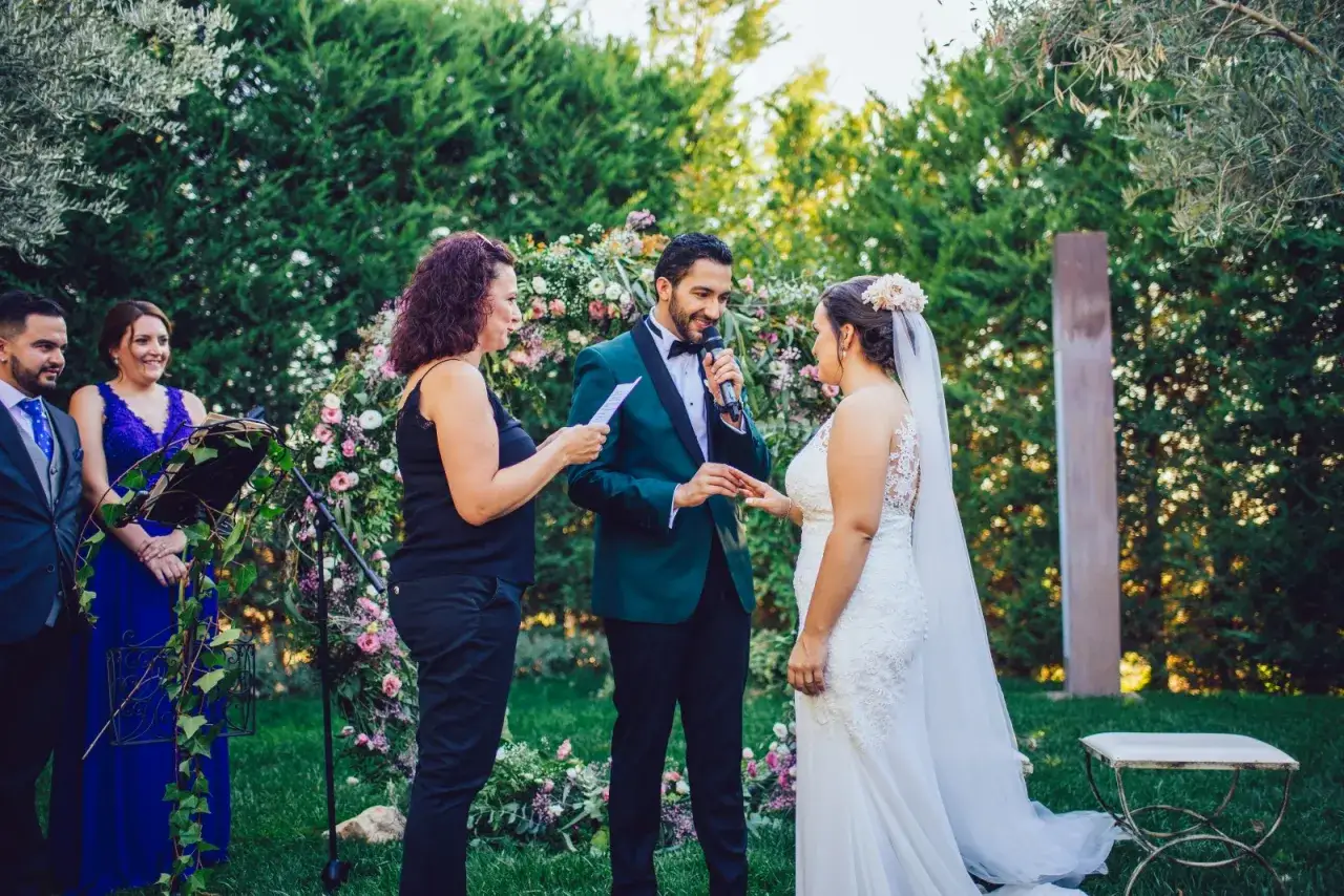 Una mujer dirige la ceremonia de boda civil. El novio, con traje verde, le pone el anillo a la novia.