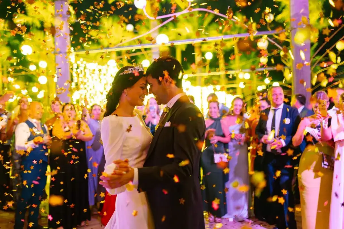 Novios bailando bajo confeti dorado, rodeados de invitados y luces cálidas. Una escena mágica que evoca diferentes tipos de boda.