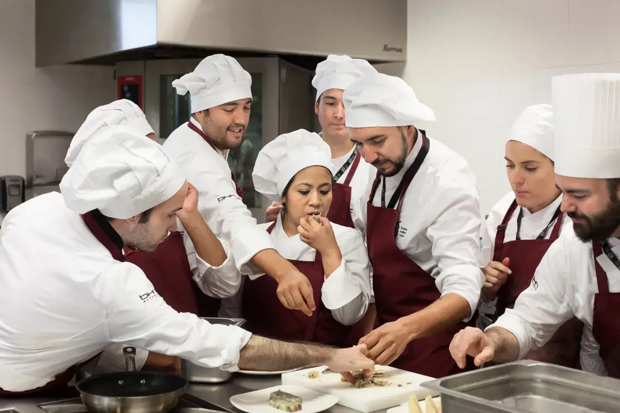 Chefs en formaci&oacute;n, con sus gorros y delantales, aprenden a preparar un plato. Uno prueba la comida, mientras otros observan y trabajan. Quiz&aacute;s est&eacute;n aprendiendo frases en euskera para la cocina.