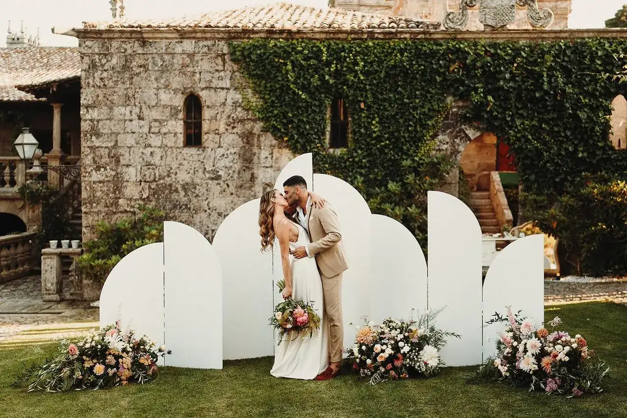 Novios besándose frente a un arco moderno con flores. Inspiración para ideas para rincones de boda.