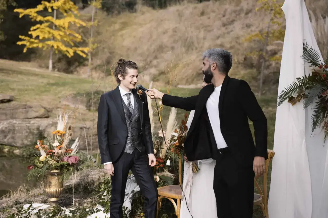 Un hombre con traje oscuro sonr&iacute;e mientras otro le acerca un micr&oacute;fono. Una sorpresa para los novios en un entorno natural.