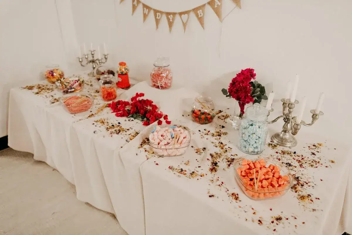 Mesa decorada con un candy bar boda casero, llena de dulces, flores rojas y candelabros.