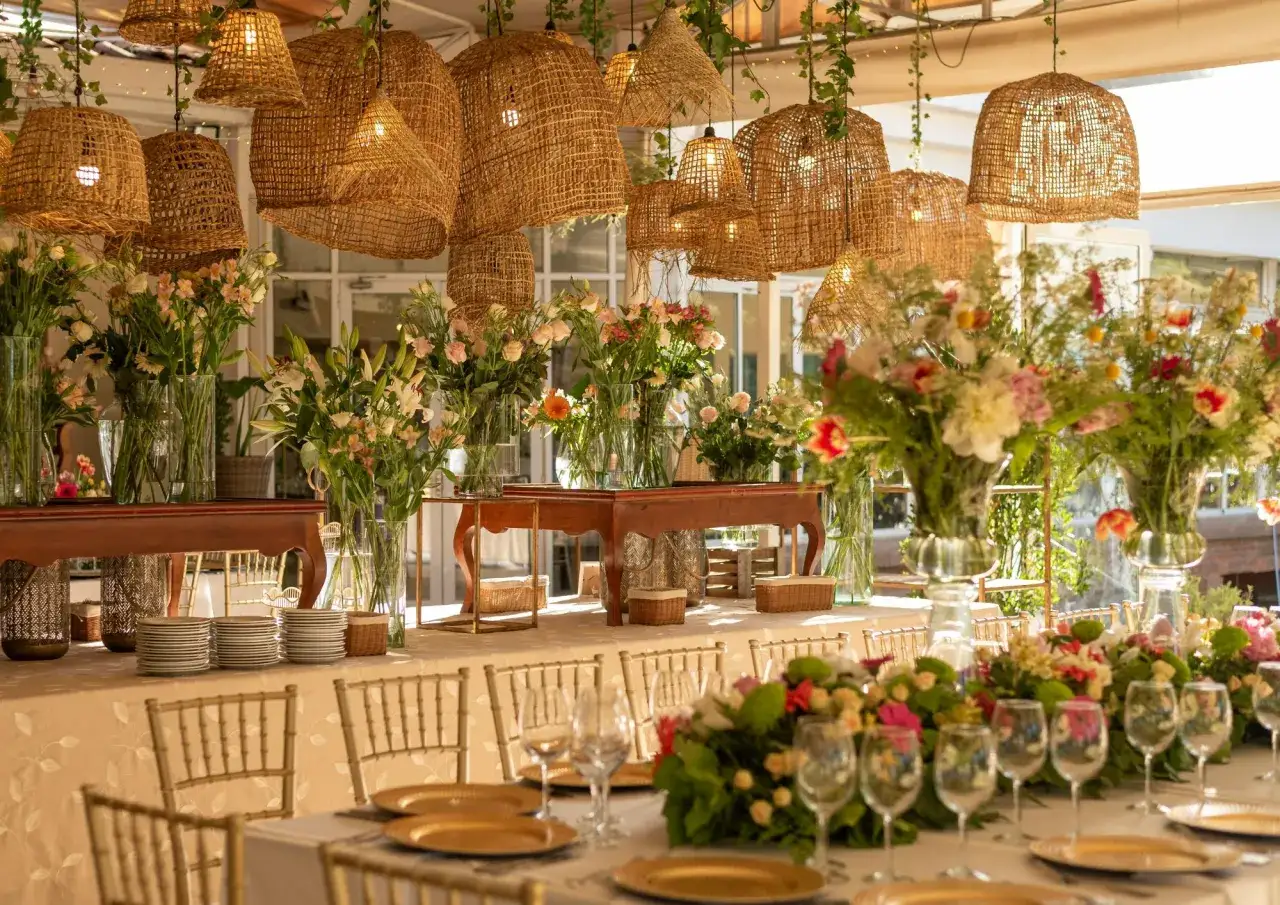 Elegante mesa puesta para un men&uacute; para bautizo, con centros de mesa florales, vajilla dorada y l&aacute;mparas de mimbre.