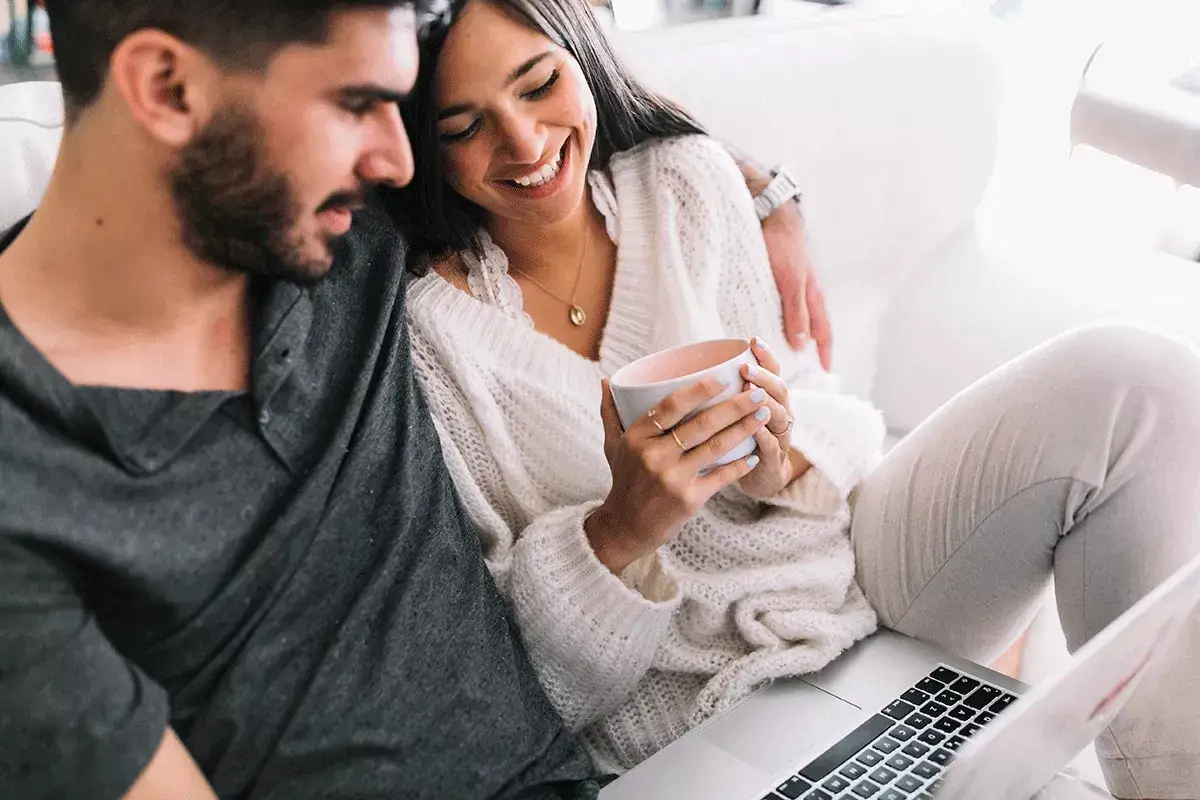 Pareja riendo en un sof&aacute;, ella con una taza, &eacute;l la abraza. Quiz&aacute;s buscando en internet 