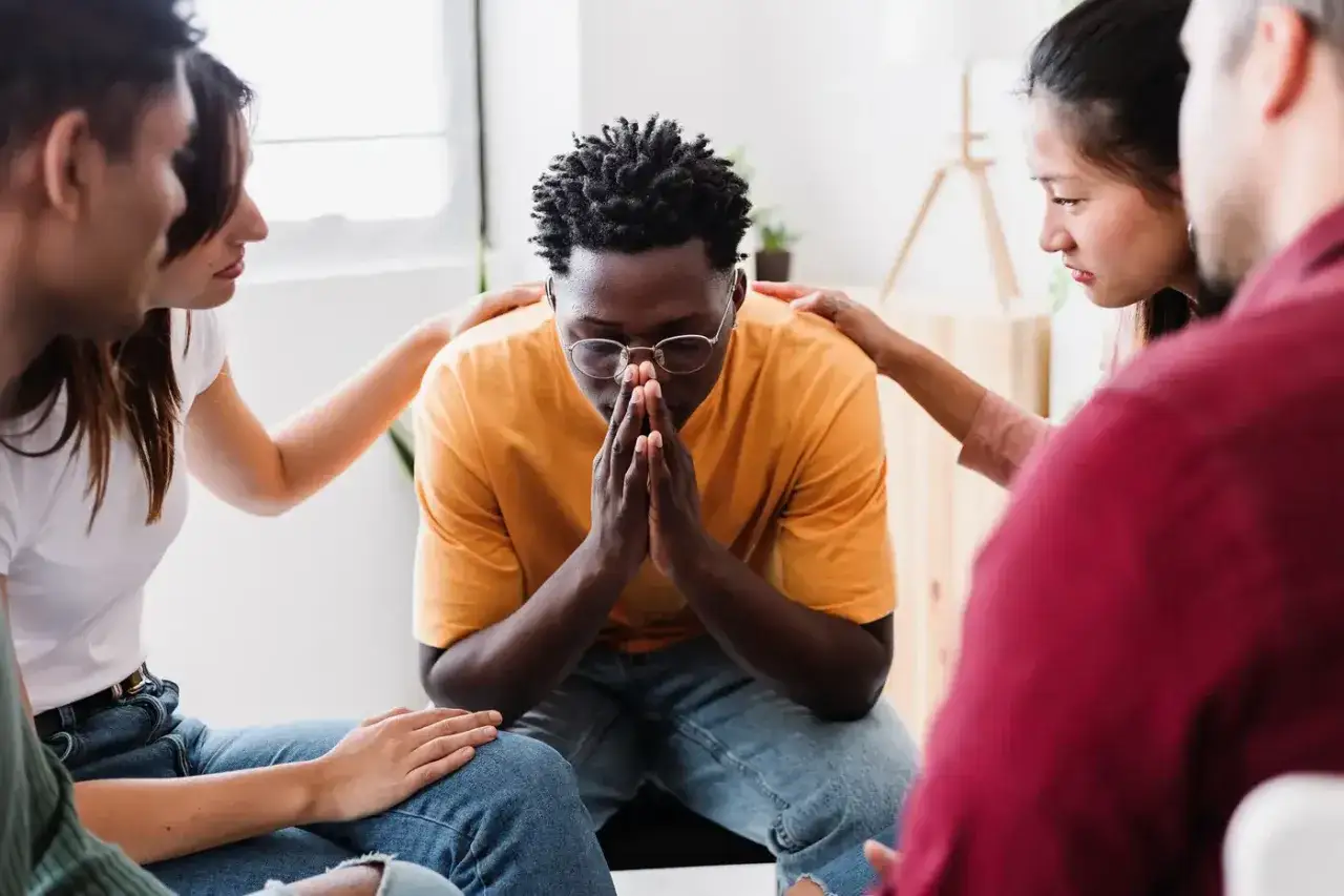 Un grupo de personas muestra apoyo a un hombre con las manos juntas, demostrando responsabilidad emocional y empatía en un momento de vulnerabilidad.