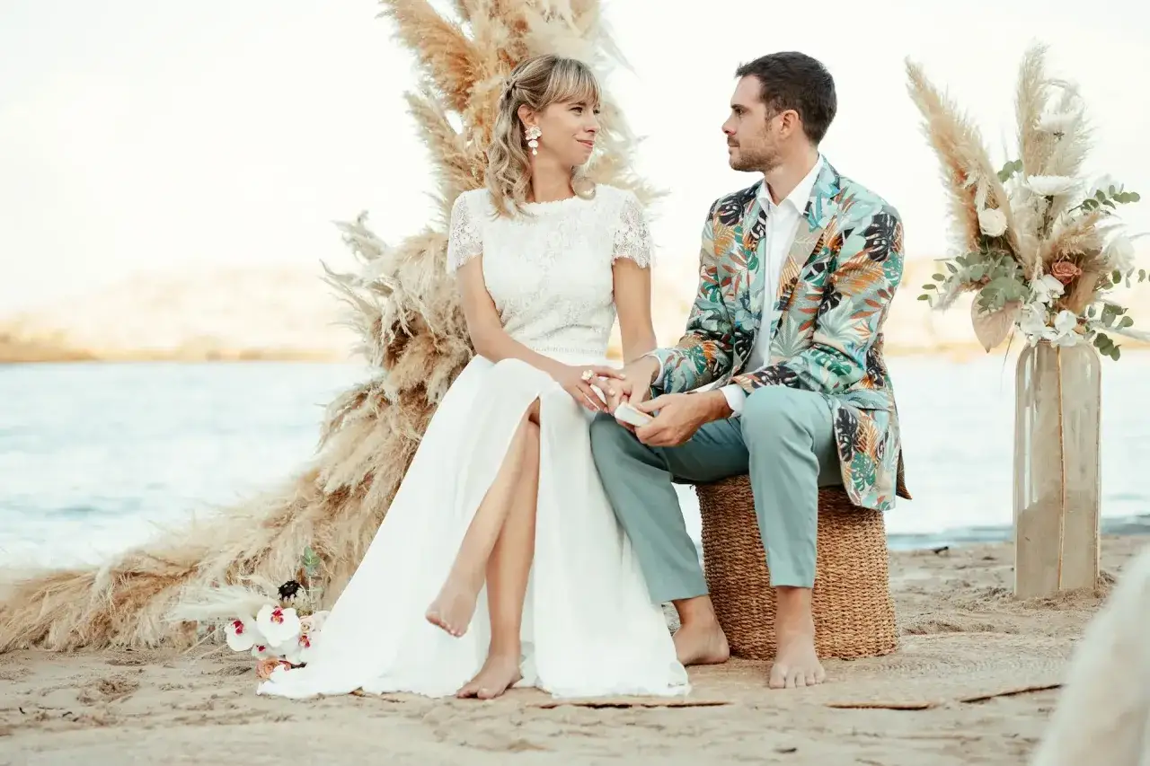 Novios en una ceremonia de boda en la playa, ella con vestido blanco, &eacute;l con chaqueta tropical.