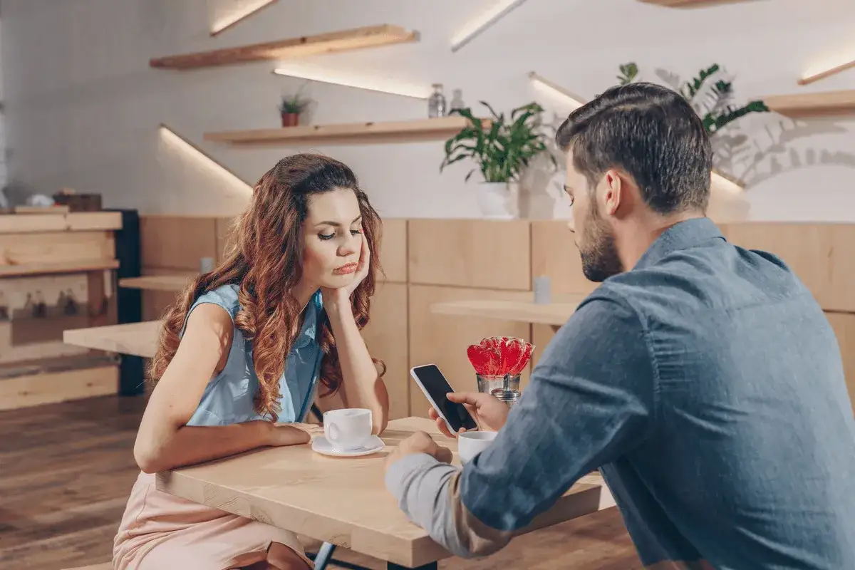 Pareja en un café, ella aburrida, él absorto en su móvil, un claro ejemplo de infidelidad emocional.