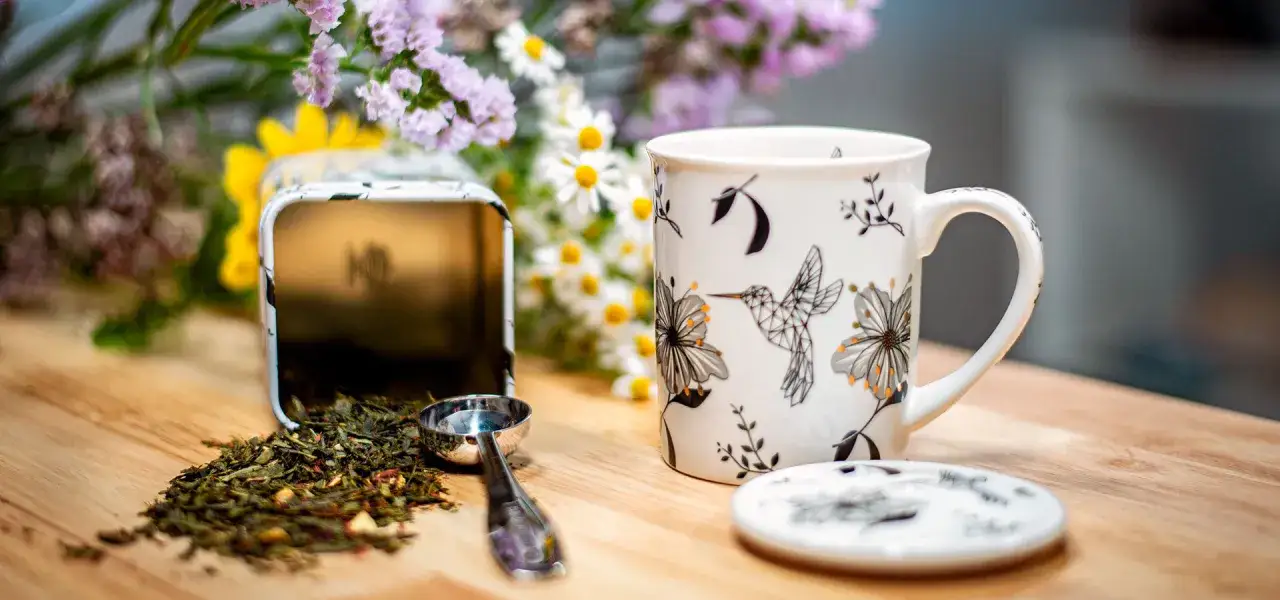 Taza decorada con colibríes y flores, junto a una lata abierta con té verde y una cuchara, ideal para disfrutar de diversos tipos de infusiones.