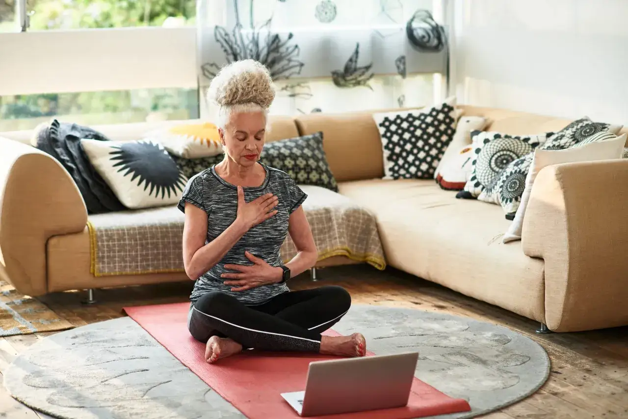 Mujer mayor practica mindfulness ejercicios en casa, sentada en un tapete rojo, con las manos en el pecho y abdomen, frente a una laptop.