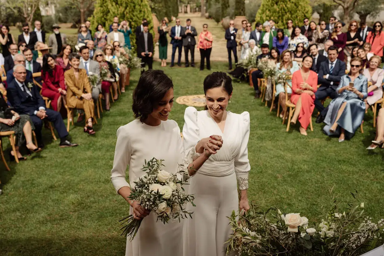 Dos novias sonr&iacute;en, cogidas de la mano, en su boda de tarde. Invitados sentados en el c&eacute;sped observan la ceremonia.