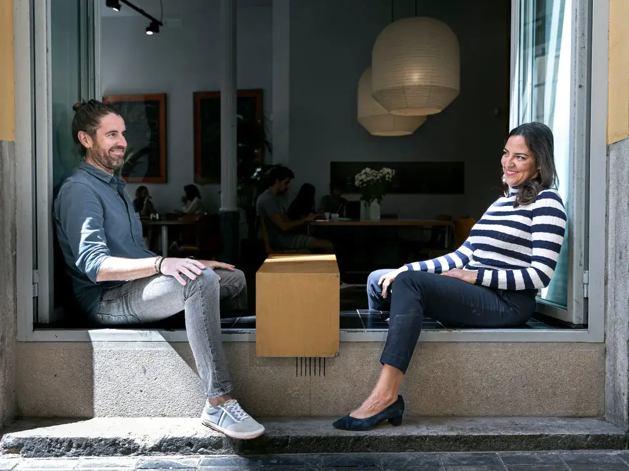 Parejas latinas conversando en la entrada de un café. Él, con coleta y barba, viste camisa azul y jeans. Ella, con cabello oscuro, lleva un jersey a rayas y pantalones oscuros.