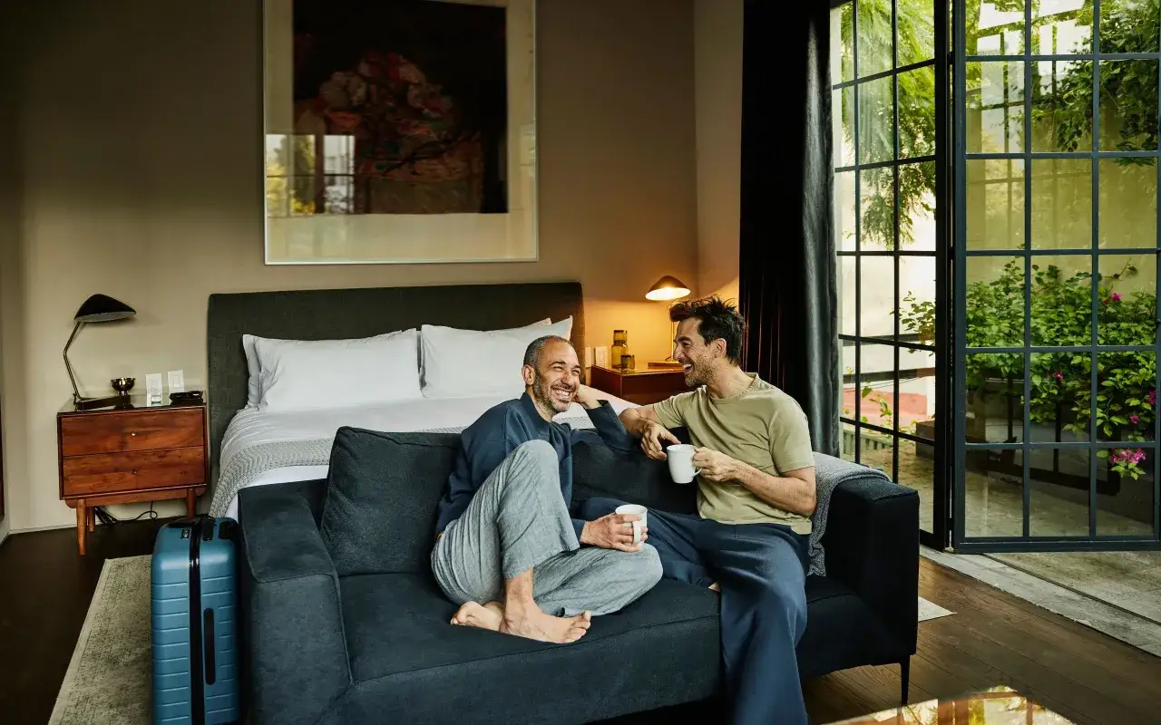 Dos hombres conversan y r&iacute;en en un sof&aacute;, tomando caf&eacute;. Uno de ellos parece preocupado, como si su pareja no lo deseara.