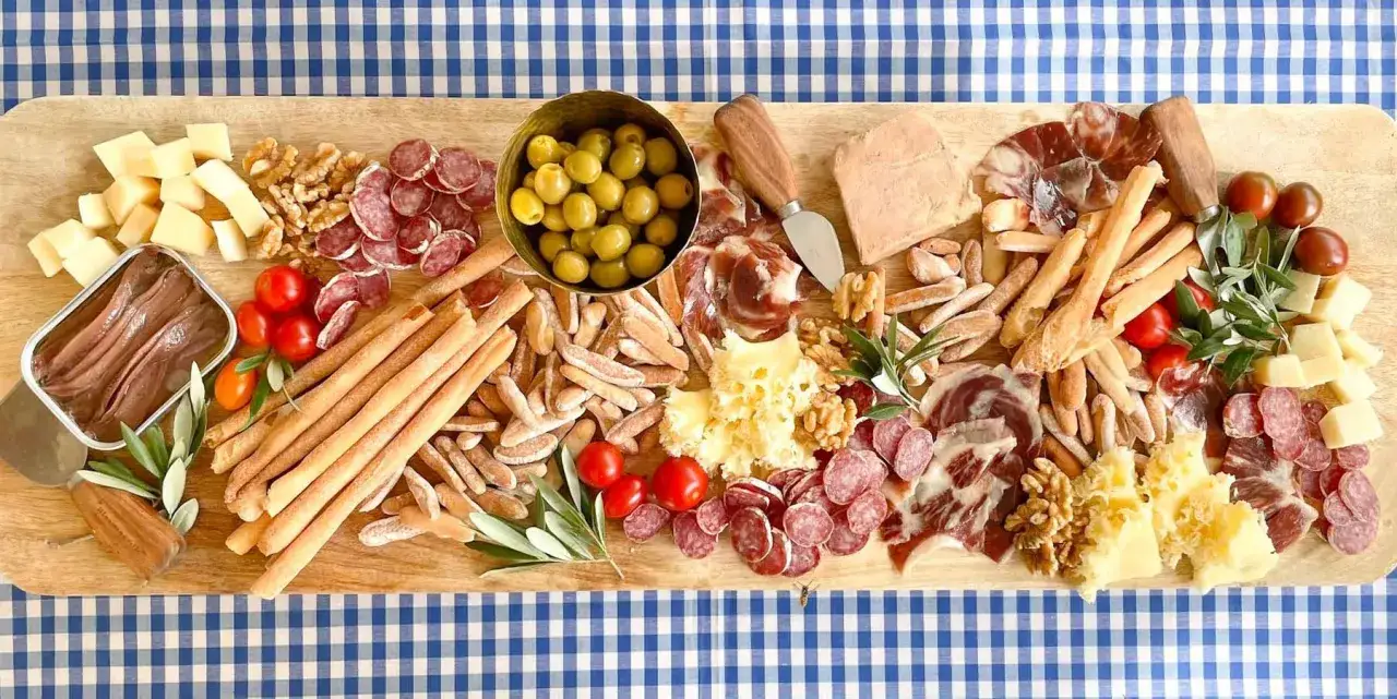 Tabla de merienda cumpleaños adultos: quesos, embutidos, aceitunas, tomates cherry, anchoas y grissini sobre tabla de madera.