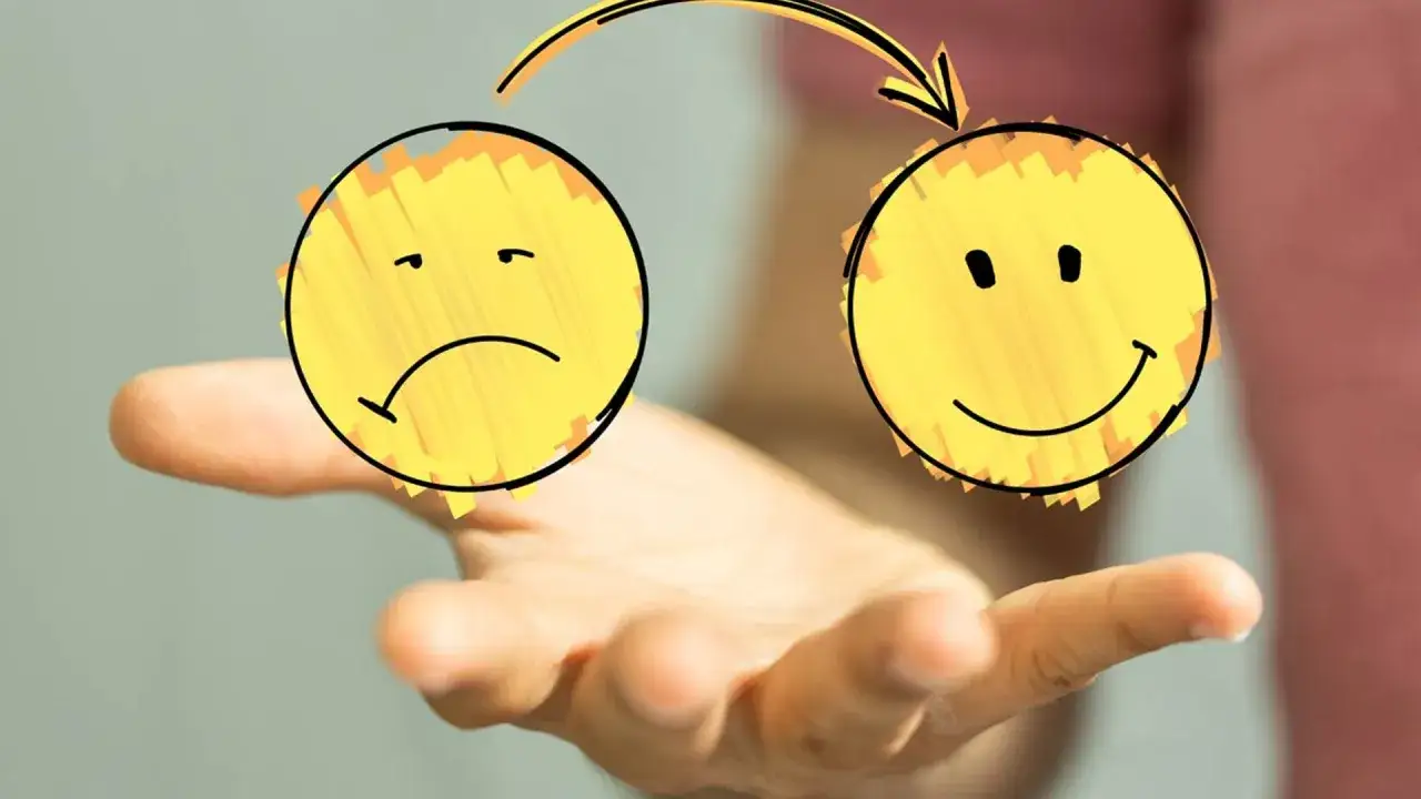 Una mano sostiene dos caritas amarillas, una triste y otra feliz, unidas por una flecha, ilustrando la labilidad emocional.