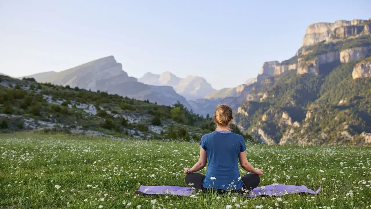 Mujer en meditaci&oacute;n en un prado de flores, disfrutando de un retiro de silencio con majestuosas monta&ntilde;as de fondo.