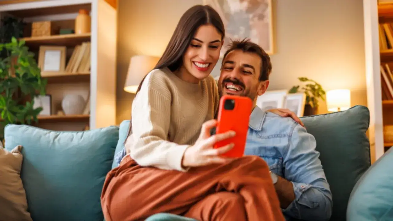 Pareja riendo mientras mira un tel&eacute;fono. Ella le hace preguntas a su novio, compartiendo un momento &iacute;ntimo y divertido en el sof&aacute;.