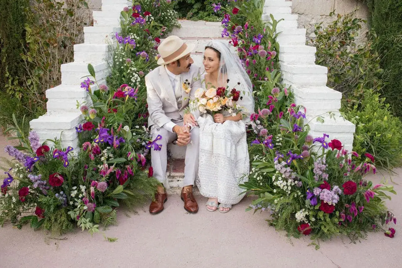 Pareja sonriente sentada en escalones blancos rodeados de flores. Ella lleva velo y vestido de encaje, él sombrero y traje. ¿Es una boda, no boda?