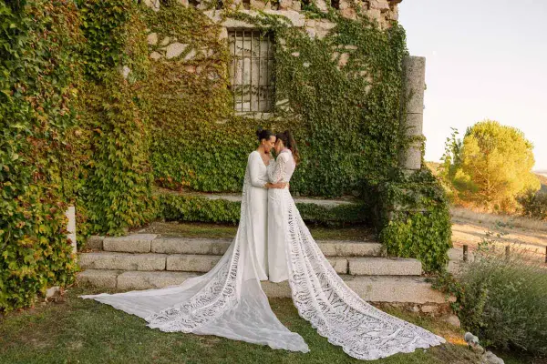 Dos novias se abrazan frente a una pared de piedra cubierta de hiedra, luciendo impresionantes ajuares de novia con velos largos y encaje.